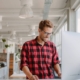 A smiling young man stands in front of his standing desk in glasses and red plaid shirt. Other office work stations are behind him.