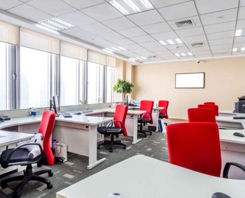 An office showcasing two parallel rows of office desks. There is a red chair and a computer at each station.