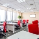 An office showcasing two parallel rows of office desks. There is a red chair and a computer at each station.