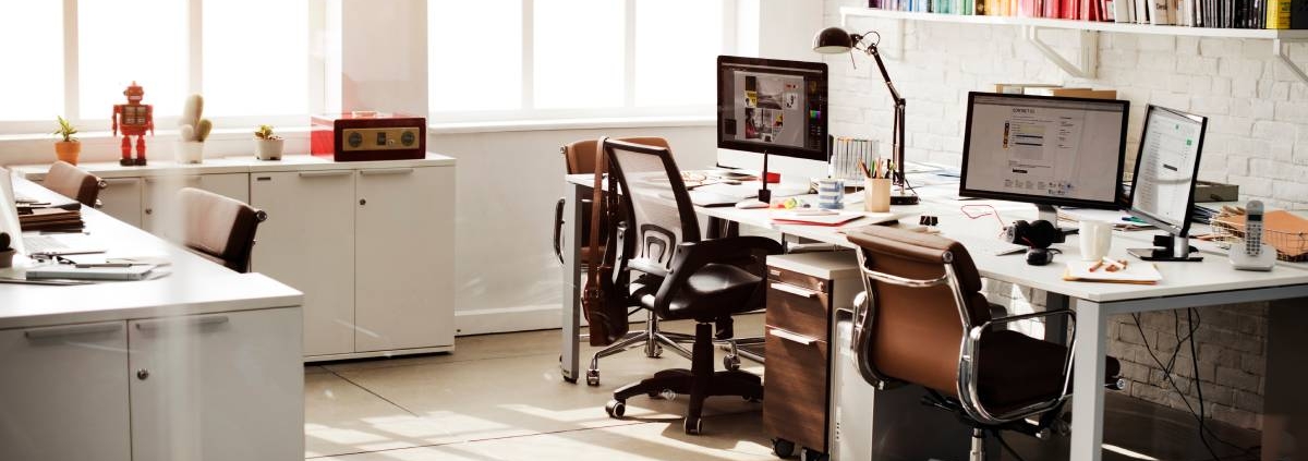 An office with two desks and brown chairs. An L-shaped workspace with cabinets is on the opposite side of the desks.