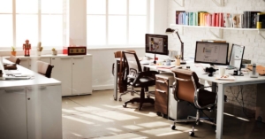 An office with two desks and brown chairs. An L-shaped workspace with cabinets is on the opposite side of the desks.