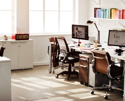 An office with two desks and brown chairs. An L-shaped workspace with cabinets is on the opposite side of the desks.