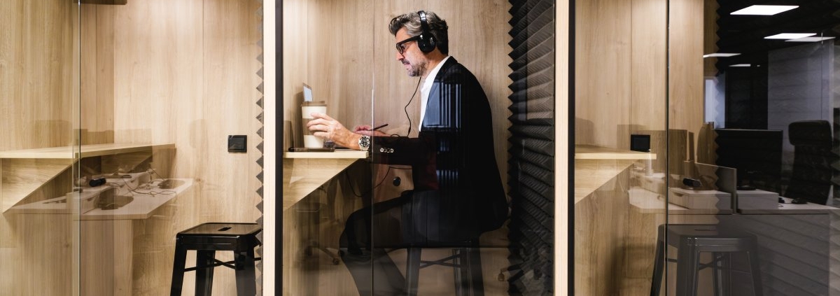 A man in a black suit sitting inside a privacy pod. He is wearing headphones and has a coffee and laptop in front of him.