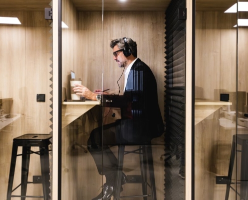 A man in a black suit sitting inside a privacy pod. He is wearing headphones and has a coffee and laptop in front of him.