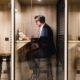 A man in a black suit sitting inside a privacy pod. He is wearing headphones and has a coffee and laptop in front of him.