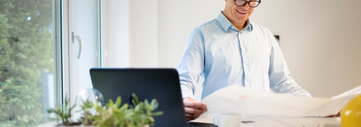 An older man wearing a blue button down stands in front of a standing desk. He smiles and holds a large paper in his hands.