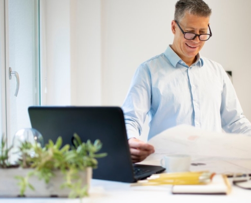 An older man wearing a blue button down stands in front of a standing desk. He smiles and holds a large paper in his hands.