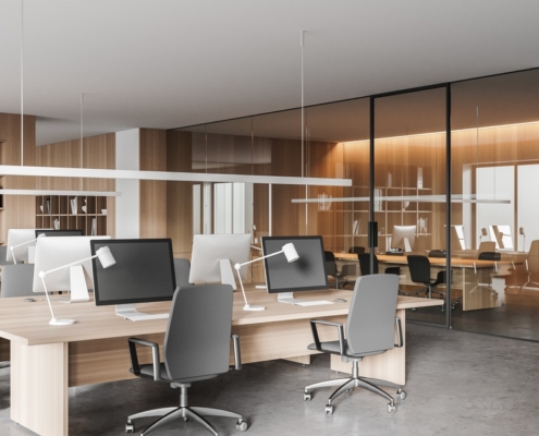 A modern office with several large desks and computers facing each other. A conference room with a table is behind the desks.