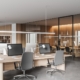 A modern office with several large desks and computers facing each other. A conference room with a table is behind the desks.
