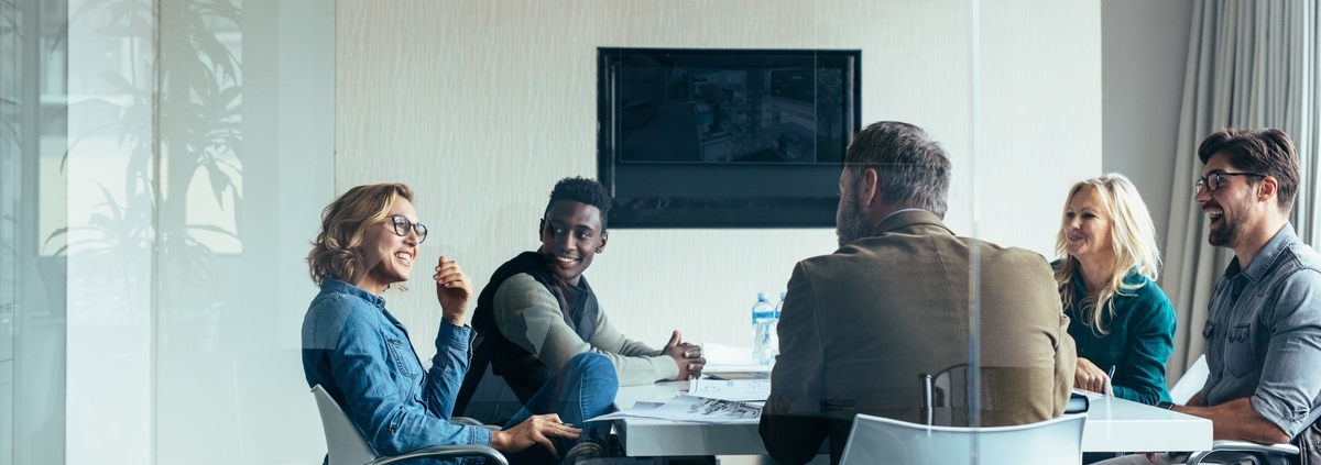 A glass-enclosed conference room with a group of coworkers sitting around a table. They are all smiling at each other.