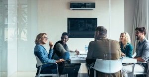 A glass-enclosed conference room with a group of coworkers sitting around a table. They are all smiling at each other.