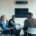 A glass-enclosed conference room with a group of coworkers sitting around a table. They are all smiling at each other.