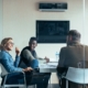 A glass-enclosed conference room with a group of coworkers sitting around a table. They are all smiling at each other.