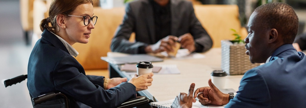 A businesswoman in a wheelchair sits at a table with two other men. She holds a coffee cup in one hand.