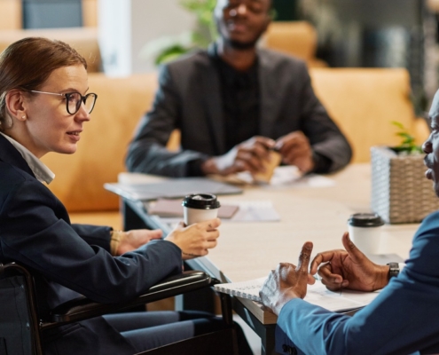 A businesswoman in a wheelchair sits at a table with two other men. She holds a coffee cup in one hand.