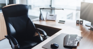 A brightly lit office space with a clean L-shaped desk. A black desk chair sits in front of the desk.