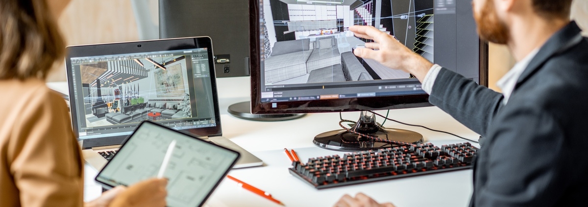 Two people sit next to each other at a computer desk. The man points to an office layout on the desktop while the woman holds a tablet.