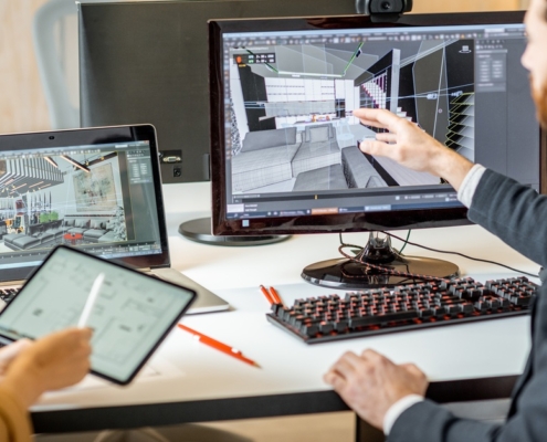 Two people sit next to each other at a computer desk. The man points to an office layout on the desktop while the woman holds a tablet.