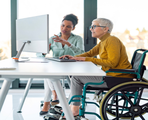 Two women sit at a desk with a monitor and keyboard. One of the women sits in a wheelchair, typing at the keyboard.