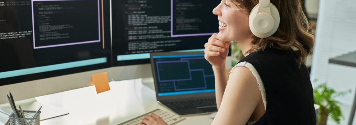 A woman sits at a desk with multiple monitors and a laptop. The surface also has a keyboard, phone, and a notebook.