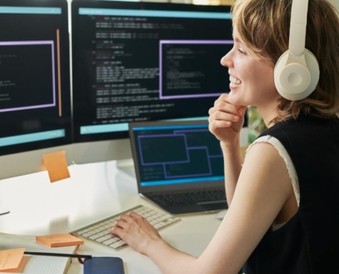 A woman sits at a desk with multiple monitors and a laptop. The surface also has a keyboard, phone, and a notebook.