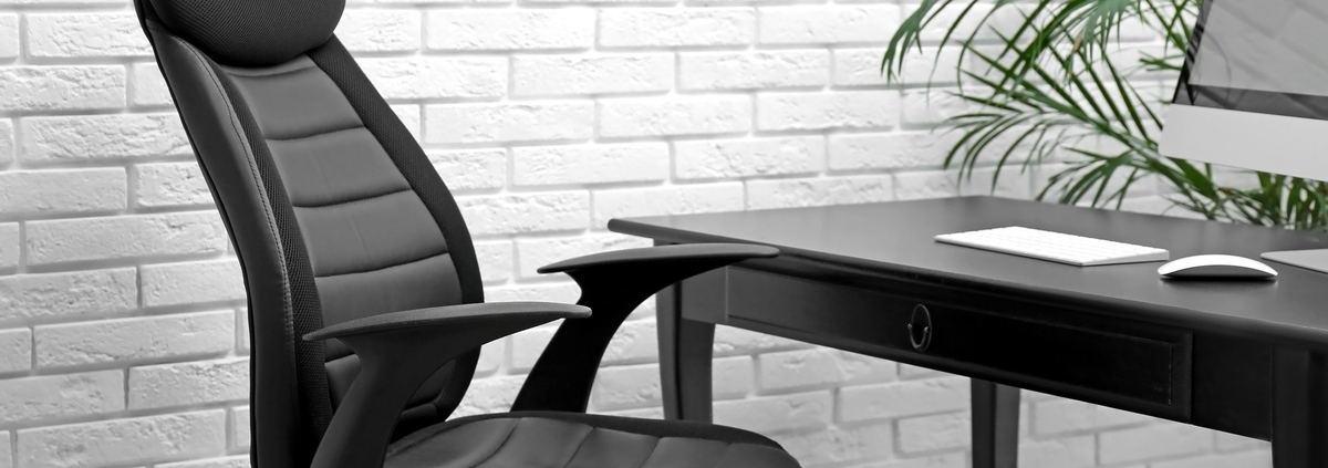 A black office chair is next to a desk. In the background is a white brick wall and a green potted plant off to the side.