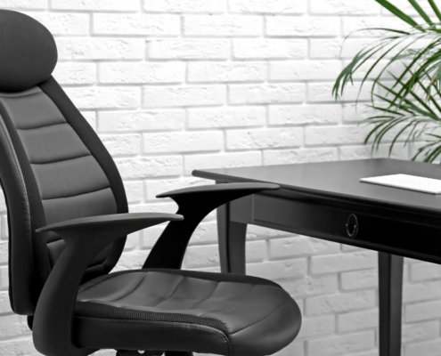 A black office chair is next to a desk. In the background is a white brick wall and a green potted plant off to the side.