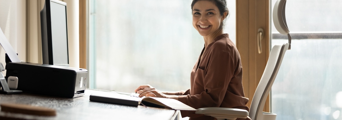A woman sits at an ergonomically designed desk. The desk surface is slightly angled, and she sits in a chair made with lumbar support.
