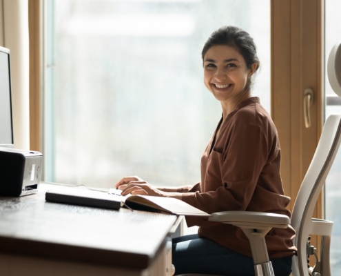 A woman sits at an ergonomically designed desk. The desk surface is slightly angled, and she sits in a chair made with lumbar support.