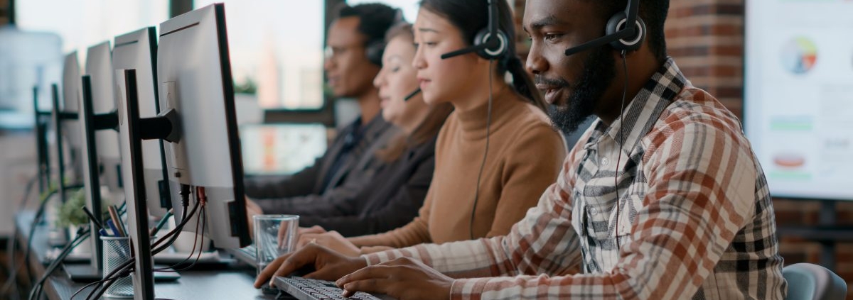 A group of co-workers sits in a line in front of computers. Each person is wearing a headset with a microphone.