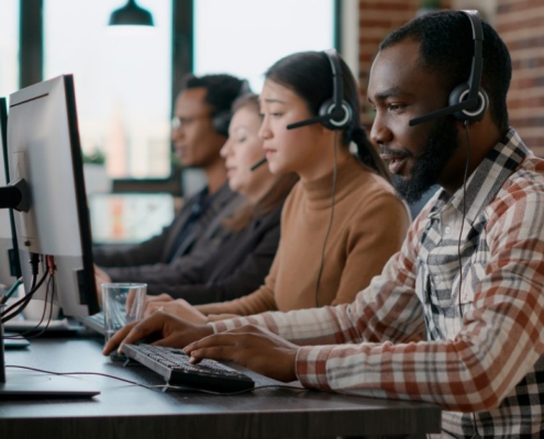 A group of co-workers sits in a line in front of computers. Each person is wearing a headset with a microphone.