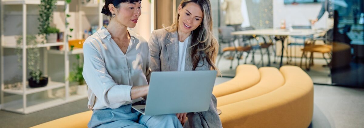 Two women are sitting on a long couch. One woman is holding a laptop while the other is looking at the screen.