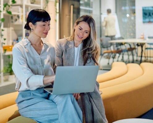 Two women are sitting on a long couch. One woman is holding a laptop while the other is looking at the screen.