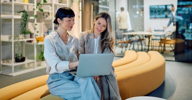 Two women are sitting on a long couch. One woman is holding a laptop while the other is looking at the screen.