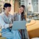 Two women are sitting on a long couch. One woman is holding a laptop while the other is looking at the screen.